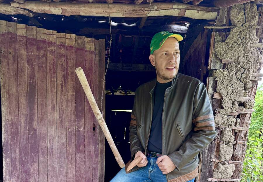Jorge Regis em pé na porta de uma casa de madeira no interior, com chapéu verde e jaqueta de couro